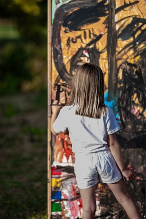 A young child with shoulder-length hair is standing and painting on a large, colorful canvas outdoors. The artwork consists of bold, abstract strokes in a variety of colors including black, orange, blue, and red. The child is wearing a white shirt and shorts, and appears to be focused on their creative task.