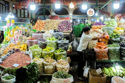 A bustling market stall filled with a diverse array of fresh fruits and vegetables. Brightly colored produce, such as carrots, lemons, limes, and purple onions, is neatly arranged in bags and baskets. A vendor is actively arranging items, contributing to the bustling atmosphere. Overhead lights illuminate the scene, highlighting the vibrant colors of the fresh produce.