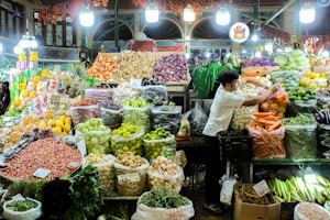 A bustling market stall filled with a diverse array of fresh fruits and vegetables. Brightly colored produce, such as carrots, lemons, limes, and purple onions, is neatly arranged in bags and baskets. A vendor is actively arranging items, contributing to the bustling atmosphere. Overhead lights illuminate the scene, highlighting the vibrant colors of the fresh produce.