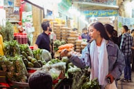 A bustling market scene with vibrant produce and goods on display. Shoppers, including a woman engaging with a vendor, convey a lively atmosphere. Stalls are filled with green vegetables, fruits, and various packages, under bright overhead lights.