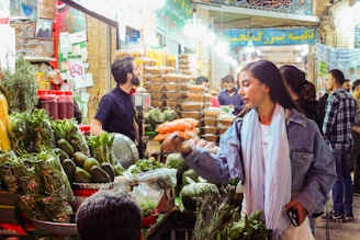 A bustling market scene with vibrant produce and goods on display. Shoppers, including a woman engaging with a vendor, convey a lively atmosphere. Stalls are filled with green vegetables, fruits, and various packages, under bright overhead lights.