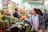 A bustling market scene with vibrant produce and goods on display. Shoppers, including a woman engaging with a vendor, convey a lively atmosphere. Stalls are filled with green vegetables, fruits, and various packages, under bright overhead lights.