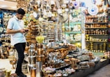 A young man in a white shirt is browsing on his phone, surrounded by a large assortment of metal cookware and decorative items in a shop. The shelves and display tables are filled with shiny, polished silver and copper plates, pots, trays, and other items, creating a vibrant and crowded visual of metallic textures and warm colors.