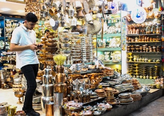 A young man in a white shirt is browsing on his phone, surrounded by a large assortment of metal cookware and decorative items in a shop. The shelves and display tables are filled with shiny, polished silver and copper plates, pots, trays, and other items, creating a vibrant and crowded visual of metallic textures and warm colors.