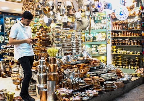 A young man in a white shirt is browsing on his phone, surrounded by a large assortment of metal cookware and decorative items in a shop. The shelves and display tables are filled with shiny, polished silver and copper plates, pots, trays, and other items, creating a vibrant and crowded visual of metallic textures and warm colors.