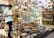 A young man in a white shirt is browsing on his phone, surrounded by a large assortment of metal cookware and decorative items in a shop. The shelves and display tables are filled with shiny, polished silver and copper plates, pots, trays, and other items, creating a vibrant and crowded visual of metallic textures and warm colors.