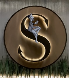 A large, illuminated letter 'S' is surrounded by a circle. A detailed, black and white illustration of a male figure with horns and a tail is seated on the 'S', holding a martini glass. The bottom of the image features tall, slender plants adding a natural element to the scene.