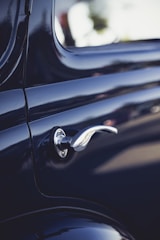 A close-up view of a vintage car door handle, showcasing its sleek metallic design against the glossy dark paint of the car body. The shiny chrome finish of the handle reflects the light, highlighting its curved and elegant shape.