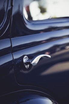 A close-up view of a vintage car door handle, showcasing its sleek metallic design against the glossy dark paint of the car body. The shiny chrome finish of the handle reflects the light, highlighting its curved and elegant shape.