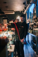 A bartender is skillfully preparing drinks behind a busy bar, surrounded by various bottles and bar tools. The atmosphere is lively with a dimly lit background, and neon lights adding a vibrant touch.