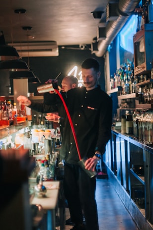 A bartender is skillfully preparing drinks behind a busy bar, surrounded by various bottles and bar tools. The atmosphere is lively with a dimly lit background, and neon lights adding a vibrant touch.