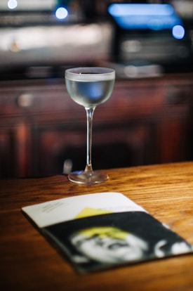 A cocktail glass filled with a clear drink is placed on a wooden surface with a blurred background of what appears to be a bar setting. In the foreground, there is an open magazine with a black and white cover featuring a partially obscured face and a yellow triangle.