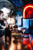 A dimly lit bar setting features a warm, yellowish lamp illuminating a collection of glassware and bottles on a wooden bar counter. In the background, colorful neon lights create a vibrant, atmospheric glow, with hints of blue and red adding to the moody ambience.