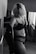 A black-and-white photo featuring a person with blonde hair in lingerie, looking down, set in an indoor location with soft light coming through a window with blinds.