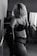 A black-and-white photo featuring a person with blonde hair in lingerie, looking down, set in an indoor location with soft light coming through a window with blinds.