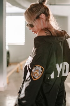 A woman wearing sunglasses and a dark jacket with a police-style patch stands in a dimly lit parking structure. Her hair is tied back, and she gazes downwards with a contemplative expression.