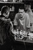 Three individuals are inside a dimly lit room playing chess on a small table. The focus is on two men engaged in the game, with one leaning over the table. Various bottles and other items can be seen on shelves in the background. The scene appears to capture a casual and concentrated atmosphere.