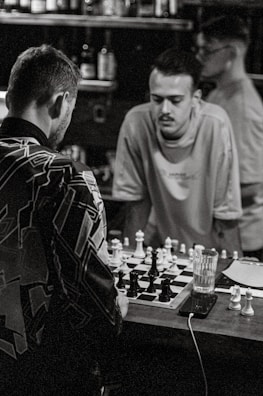 Three individuals are inside a dimly lit room playing chess on a small table. The focus is on two men engaged in the game, with one leaning over the table. Various bottles and other items can be seen on shelves in the background. The scene appears to capture a casual and concentrated atmosphere.
