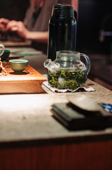 A glass teapot with green tea leaves steeping inside is placed on a cloth coaster on a wooden surface. There is a black thermos behind the teapot and a small green teacup on a wooden tray. The background is softly blurred, suggesting an indoor, warm, and cozy setting, possibly in a tea house or cafe.
