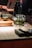A glass teapot with green tea leaves steeping inside is placed on a cloth coaster on a wooden surface. There is a black thermos behind the teapot and a small green teacup on a wooden tray. The background is softly blurred, suggesting an indoor, warm, and cozy setting, possibly in a tea house or cafe.