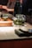 A glass teapot with green tea leaves steeping inside is placed on a cloth coaster on a wooden surface. There is a black thermos behind the teapot and a small green teacup on a wooden tray. The background is softly blurred, suggesting an indoor, warm, and cozy setting, possibly in a tea house or cafe.