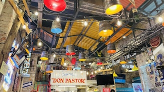 An indoor setting with colorful industrial-style lighting fixtures, featuring brightly colored buckets hanging from the ceiling and various signs and decorative elements. The walls and roof showcase a rustic warehouse aesthetic with exposed beams and ambient neon lighting.