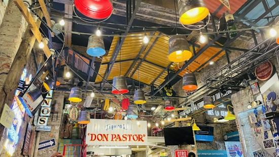 An indoor setting with colorful industrial-style lighting fixtures, featuring brightly colored buckets hanging from the ceiling and various signs and decorative elements. The walls and roof showcase a rustic warehouse aesthetic with exposed beams and ambient neon lighting.