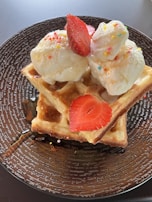 A delicious dessert featuring two scoops of ice cream adorned with colorful sprinkles, placed on top of golden-brown waffles. Fresh strawberry slices are laid atop the ice cream. A drizzle of syrup can be seen on the waffles and the textured brown plate underneath.