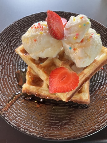 A delicious dessert featuring two scoops of ice cream adorned with colorful sprinkles, placed on top of golden-brown waffles. Fresh strawberry slices are laid atop the ice cream. A drizzle of syrup can be seen on the waffles and the textured brown plate underneath.