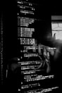Lines of computer code are displayed on a screen in a dark setting, with a faint reflection of a person taking a picture in the background.