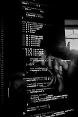 Lines of computer code are displayed on a screen in a dark setting, with a faint reflection of a person taking a picture in the background.