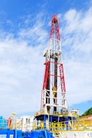 A large oil drilling rig with a towering red and white structure stands against a sky with scattered clouds. The base of the rig features industrial equipment and pipes, predominantly in blue and yellow. There is construction signage and caution symbols visible, indicating a work site.