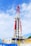 A large oil drilling rig with a towering red and white structure stands against a sky with scattered clouds. The base of the rig features industrial equipment and pipes, predominantly in blue and yellow. There is construction signage and caution symbols visible, indicating a work site.