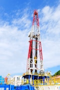 A large oil drilling rig with a towering red and white structure stands against a sky with scattered clouds. The base of the rig features industrial equipment and pipes, predominantly in blue and yellow. There is construction signage and caution symbols visible, indicating a work site.