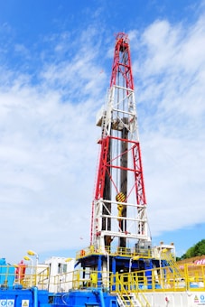 A large oil drilling rig with a towering red and white structure stands against a sky with scattered clouds. The base of the rig features industrial equipment and pipes, predominantly in blue and yellow. There is construction signage and caution symbols visible, indicating a work site.
