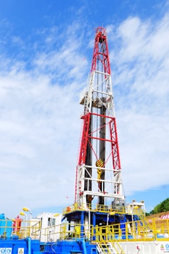 A large oil drilling rig with a towering red and white structure stands against a sky with scattered clouds. The base of the rig features industrial equipment and pipes, predominantly in blue and yellow. There is construction signage and caution symbols visible, indicating a work site.