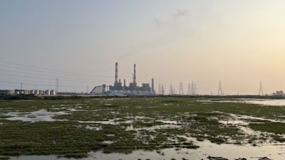 An industrial facility with multiple large chimneys emitting smoke is situated in the distance. Surrounding the facility are power lines and electricity pylons. In the foreground, there is a marshy area with patches of grass and water reflecting the sky.