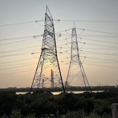 Large metal electricity pylons stand in the foreground, crisscrossed with power lines. Behind them, the sun is setting, casting a warm glow in the sky. A body of water sits in front of a distant industrial complex.