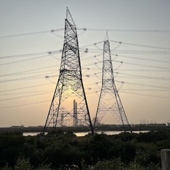 Large metal electricity pylons stand in the foreground, crisscrossed with power lines. Behind them, the sun is setting, casting a warm glow in the sky. A body of water sits in front of a distant industrial complex.
