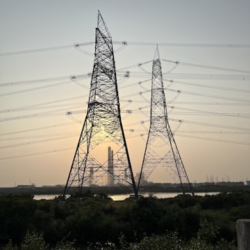 Large metal electricity pylons stand in the foreground, crisscrossed with power lines. Behind them, the sun is setting, casting a warm glow in the sky. A body of water sits in front of a distant industrial complex.