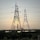 Large metal electricity pylons stand in the foreground, crisscrossed with power lines. Behind them, the sun is setting, casting a warm glow in the sky. A body of water sits in front of a distant industrial complex.