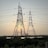 Large metal electricity pylons stand in the foreground, crisscrossed with power lines. Behind them, the sun is setting, casting a warm glow in the sky. A body of water sits in front of a distant industrial complex.