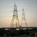 Large metal electricity pylons stand in the foreground, crisscrossed with power lines. Behind them, the sun is setting, casting a warm glow in the sky. A body of water sits in front of a distant industrial complex.