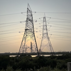 Large metal electricity pylons stand in the foreground, crisscrossed with power lines. Behind them, the sun is setting, casting a warm glow in the sky. A body of water sits in front of a distant industrial complex.