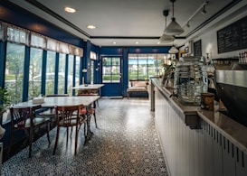 A cozy caf&eacute; with large windows allowing natural light to fill the space. The interior features blue and white decor, with a long counter displaying a large glass water dispenser and decorative items. There's a patterned tile floor, white tables, dark wooden chairs, and a coffee machine visible behind the counter. Outside, greenery can be seen through the windows.