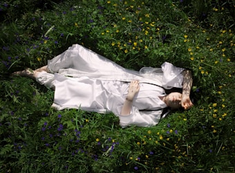 A person in a white robe lies peacefully on lush, green grass surrounded by small yellow and purple flowers. Their arm is raised, casting a shadow across their face. The scene suggests tranquility and harmony with nature.