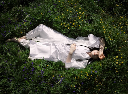 A person in a white robe lies peacefully on lush, green grass surrounded by small yellow and purple flowers. Their arm is raised, casting a shadow across their face. The scene suggests tranquility and harmony with nature.