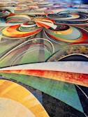 A vibrant and intricate carpet design featuring a complex arrangement of swirling patterns and arcs in various colors including red, orange, yellow, green, and blue. The design also incorporates dotted lines and layered textures, creating a dynamic and visually stimulating effect.