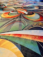 A vibrant and intricate carpet design featuring a complex arrangement of swirling patterns and arcs in various colors including red, orange, yellow, green, and blue. The design also incorporates dotted lines and layered textures, creating a dynamic and visually stimulating effect.