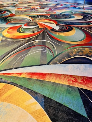 A vibrant and intricate carpet design featuring a complex arrangement of swirling patterns and arcs in various colors including red, orange, yellow, green, and blue. The design also incorporates dotted lines and layered textures, creating a dynamic and visually stimulating effect.